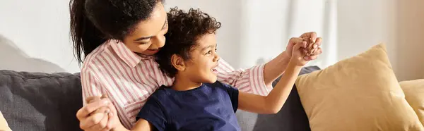 Jolly african american mother holding her little cute son and sitting on sofa in living room, banner — Stock Photo