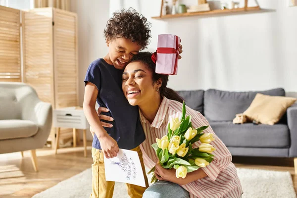 Jolly african american woman hugging her son with postcard present and flowers, Mothers day — Stock Photo