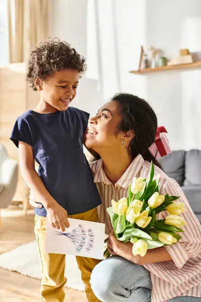 Jolly african american woman with postcard and flowers looking happily at her son, Mothers day — Stock Photo