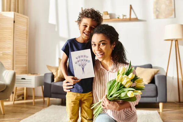 Joyful african american woman with postcard and flowers hugging her son and looking at camera — Stock Photo