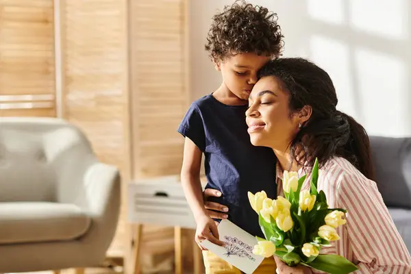 Joyous african american woman hugging her son and holding postcard and tulips, modern parenting — Stock Photo