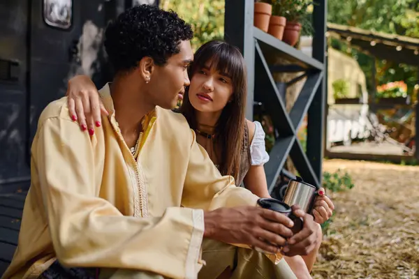 A man and woman, an interracial couple, sit peacefully in a camper van, enjoying each others company while holding cups. — Stock Photo