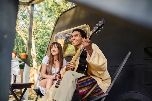 Ein Mann spielt eine Gitarre, während er neben einer Frau sitzt und in einer natürlichen Umgebung am Lagerfeuer eine harmonische Melodie erzeugt. — Stockfoto