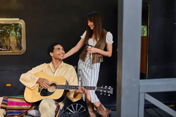 A man playing a guitar next to a woman, creating music in a natural setting during a romantic camper van getaway. — Stock Photo