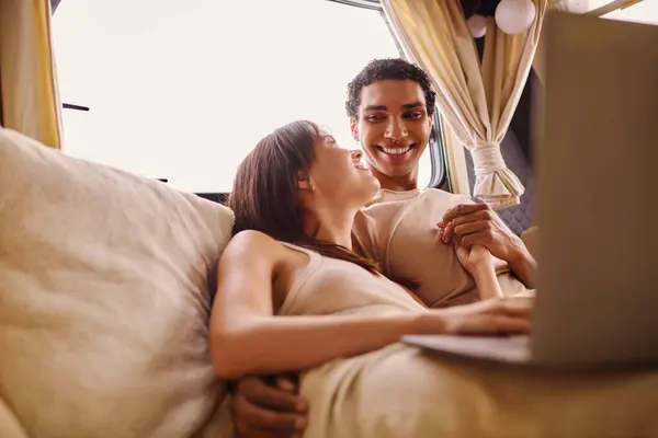 Interracial man and woman are lying comfortably on a couch, using a laptop together during a cozy evening at home. — Stock Photo