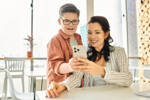 Cheerful boy with Down syndrome spending time with his beautiful mother in cafe, holding smartphone — Stock Photo