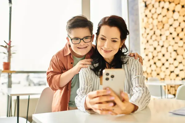 Cheerful boy with Down syndrome spending time with his beautiful mother in cafe, holding smartphone — Stock Photo
