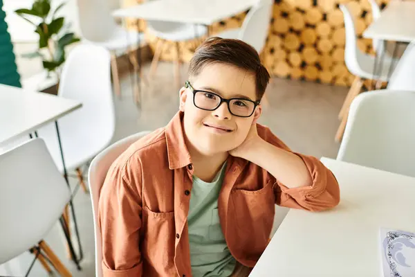 Alegre inclusivo preadolescente chico con síndrome de Down con gafas sentado en la cafetería y sonriendo a la cámara - foto de stock