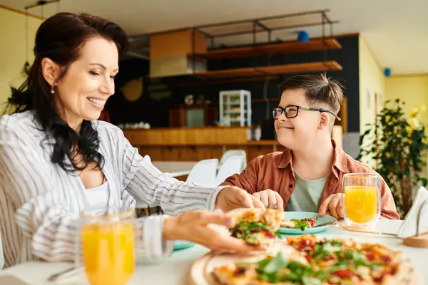 Alegre madre comer pizza y beber jugo con su inclusivo lindo hijo con síndrome de Down - foto de stock