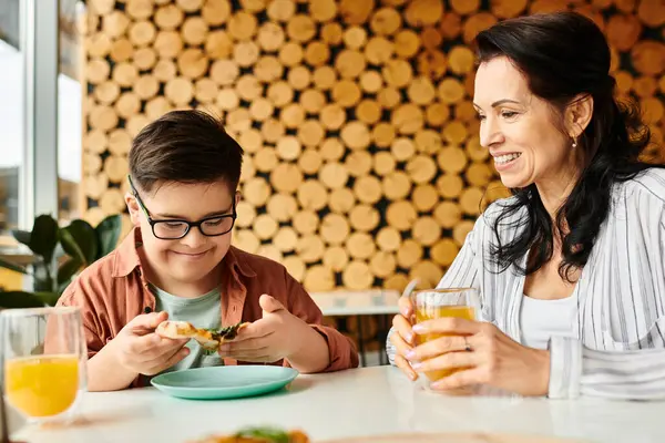 Веселая мать ест пиццу и пьет сок с ее включительно милый сын с синдромом Дауна — стоковое фото