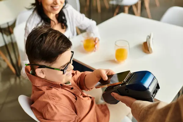 Adorable inclusive boy with Down syndrome paying with smartphone in cafe near his jolly mother — Stock Photo