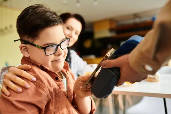 Adorable niño inclusivo con síndrome de Down pagando con teléfono inteligente en la cafetería cerca de su alegre madre - foto de stock