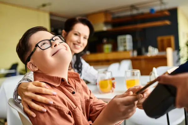 Entzückender inklusiver Junge mit Down-Syndrom bezahlt mit Smartphone im Café in der Nähe seiner fröhlichen Mutter — Stockfoto