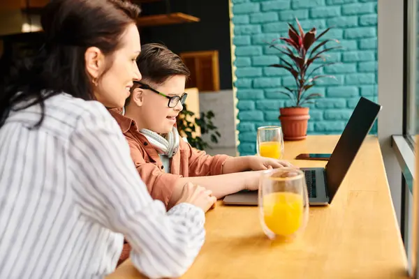 Веселый инклюзивный мальчик с синдромом Дауна с наушниками, играющий на ноутбуке рядом с матерью в кафе — стоковое фото