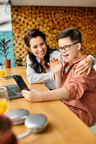 Весело включенный мальчик с синдромом Дауна машет перед камерой телефона на видео-звонок рядом с его матерью в кафе — стоковое фото