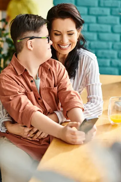 Веселый инклюзивный мальчик с синдромом Дауна смотрит на телефон рядом со своей веселой матерью в кафе — стоковое фото
