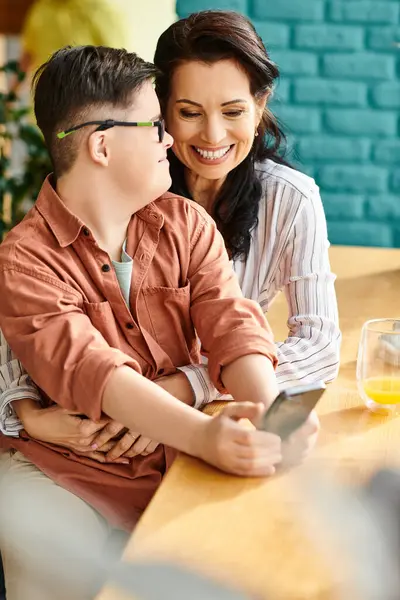 Joyous inclusive boy with Down syndrome looking at phone next to his cheerful mother in cafe — Stock Photo