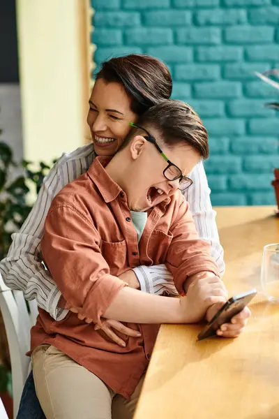 Joyous inclusive boy with Down syndrome looking at phone next to his cheerful mother in cafe — Stock Photo