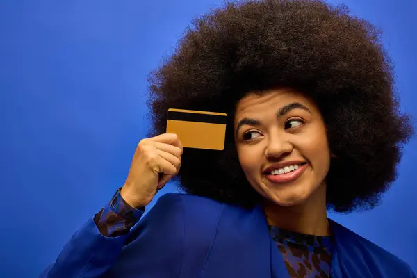 Elegante mujer afroamericana con el pelo rizado sosteniendo una tarjeta de crédito en un vibrante telón de fondo. - foto de stock