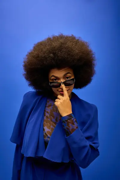 Stylish African American woman posing in trendy blue coat and glasses on vibrant backdrop. — Stock Photo
