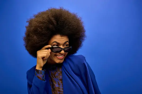 African American woman with curly hairdohair holding stylish sunglasses indoors against a vibrant backdrop. — Stock Photo