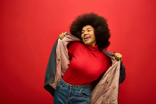 Stylish African American woman with curly hairdohair, holding jacket, posing against vibrant backdrop. — Stock Photo