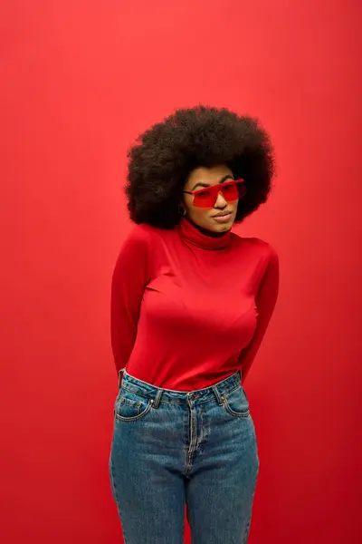 African American woman posing in trendy red shirt and jeans against vibrant backdrop. — Stock Photo