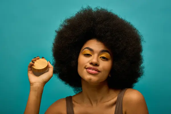 Stylish African American woman with curly hairdoholding cream on vibrant backdrop. — Stock Photo