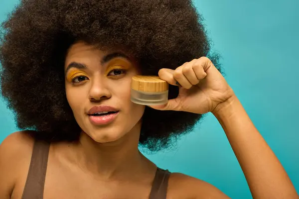Elegante mujer afroamericana con el pelo rizado posando con un frasco de maquillaje. - foto de stock