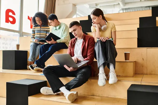 A multicultural group of students sit on steps, working together on a laptop in an educational setting. — Stock Photo