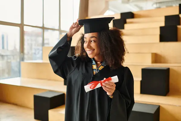 A young African American woman stands proudly in a graduation cap and gown, symbolizing academic success and accomplishments. — Stock Photo