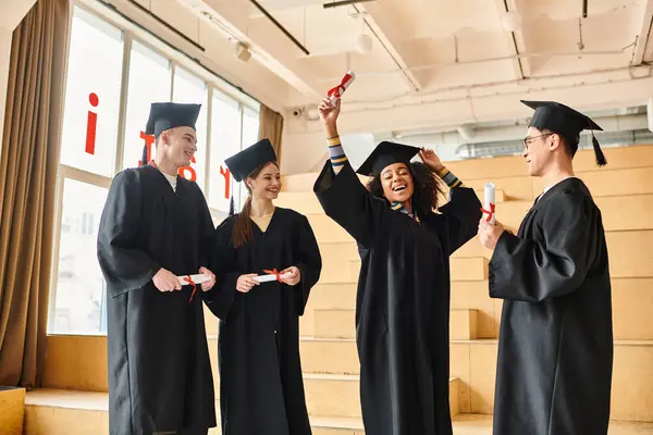 Grupo de estudiantes multiculturales en trajes de graduación levantan sus manos alegremente. - foto de stock