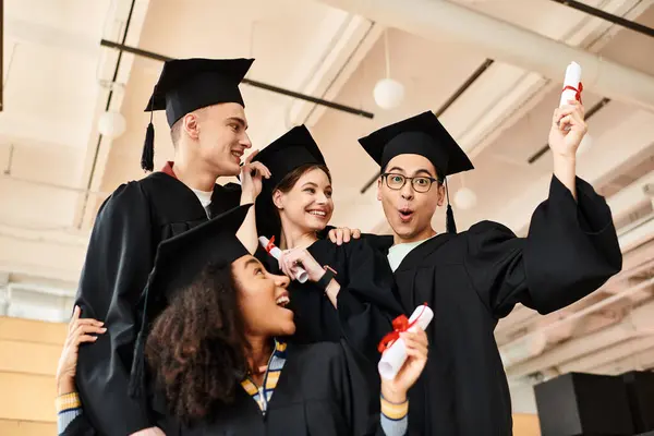 Diverso grupo de estudiantes en vestidos de graduación y gorras felizmente tomando una selfie juntos. - foto de stock