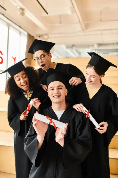 Un grupo diverso de estudiantes en trajes de graduación y gorras académicas posando felizmente para una foto en el interior. - foto de stock
