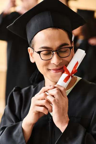 Asian man wearing a graduation cap and gown smiling while holding a diploma in his hand. — Stock Photo