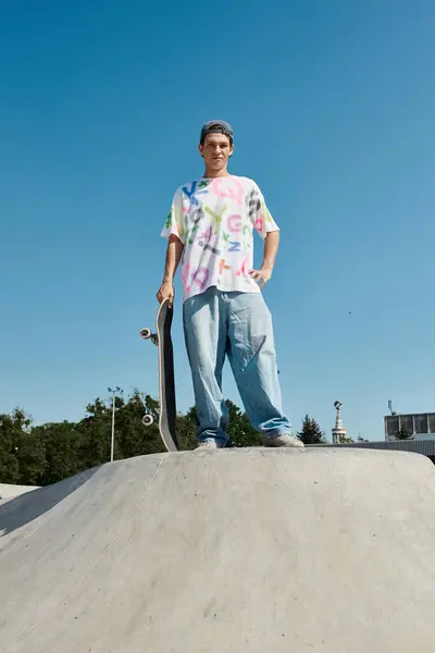 A young skater confidently holds a skateboard on top of a ramp in a vibrant outdoor skate park on a sunny summer day. — Stock Photo