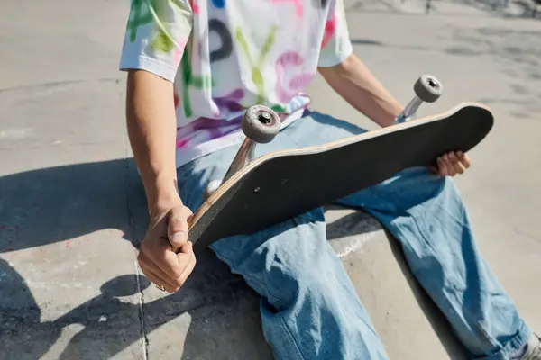 Um jovem senta-se em uma laje de cimento, segurando casualmente um skate, aproveitando o dia de verão em um parque de skate ao ar livre. — Fotografia de Stock