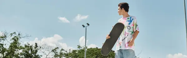 A young man holds a skateboard in his hand at a skate park outdoors on a summer day. — Stock Photo