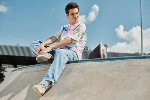 Ein junger Mann sitzt an einem sonnigen Sommertag selbstbewusst auf einer Skateboard-Rampe in einem lebhaften Outdoor-Skatepark. — Stockfoto