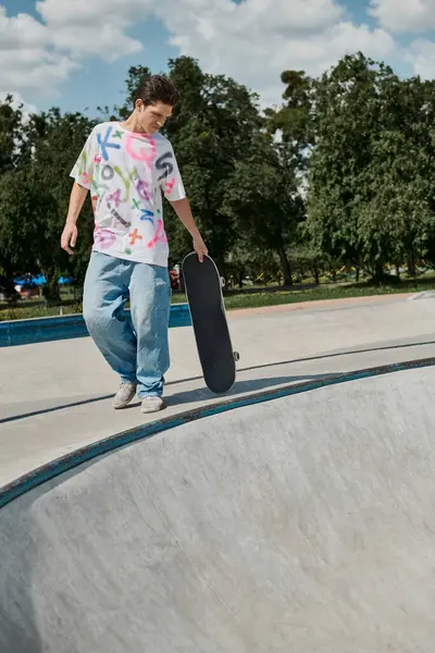 Молодой человек уверенно держит скейтборд, стоя на рампе скейтборда в открытом скейтпарке в солнечный летний день. — стоковое фото