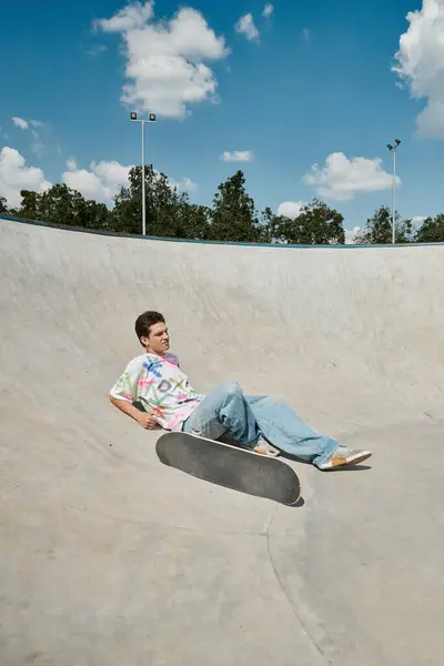 Un joven patinador descansando cerca del monopatín en una rampa en un bullicioso parque de skate al aire libre en un soleado día de verano. - foto de stock