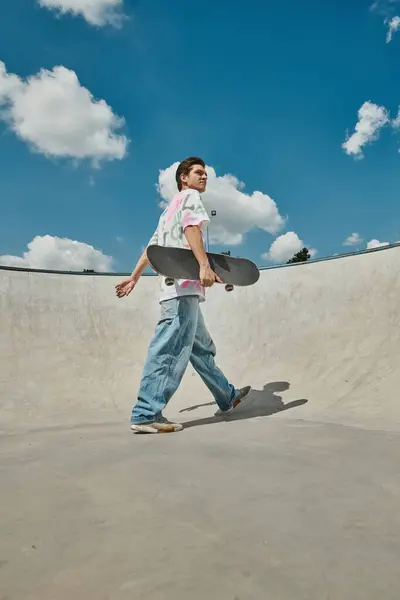 Un jeune homme marche tranquillement avec une planche à roulettes à la main, respirant une ambiance fraîche et insouciante dans un cadre de skate park d'été. — Photo de stock