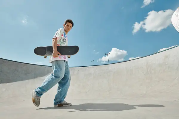 Ein junger Mann läuft selbstbewusst in einem Skatepark und hält sein Skateboard in der prallen Sonne. — Stockfoto
