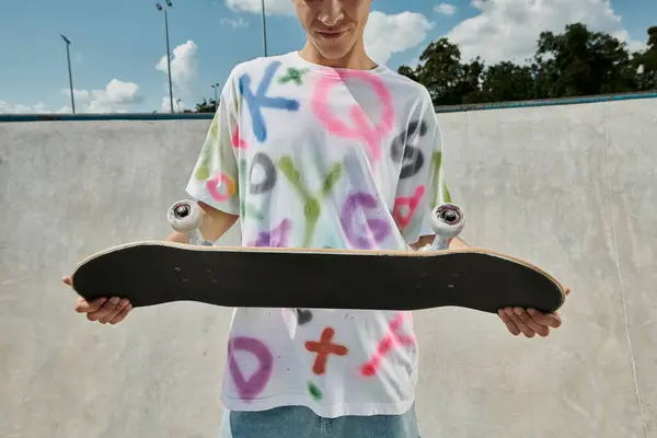 Un jeune homme tient en toute confiance une planche à roulettes dans ses mains dans un skate park par une journée d'été ensoleillée. — Photo de stock