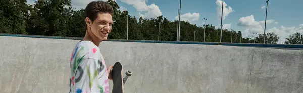 Молодий чоловік, прикрашений яскравою сорочкою, радісно тримає скейтборд у галасливому ковзанярському парку в сонячний день. — стокове фото