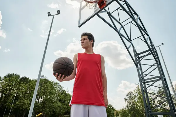 Un joven con una camisa roja se levanta al aire libre, sosteniendo una pelota de baloncesto en su mano en un día soleado de verano.. - foto de stock