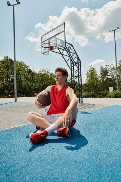 Un joven jugador de baloncesto se sienta en la cancha, pensando profundamente, sosteniendo una pelota de baloncesto en sus manos en un día soleado de verano.. - foto de stock