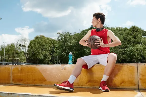 Un jeune joueur de basket assis sur une corniche, tenant habilement un ballon de basket. — Photo de stock
