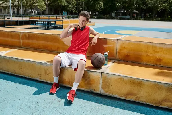 Un hombre con una camisa roja y pantalones cortos blancos se sienta en las escaleras con una pelota de baloncesto, mostrando su pasión por el juego en un entorno al aire libre. - foto de stock