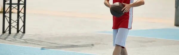 Um jovem jogador de basquete fica em cima de uma quadra de basquete, segurando com confiança uma bola em um dia ensolarado de verão. — Fotografia de Stock
